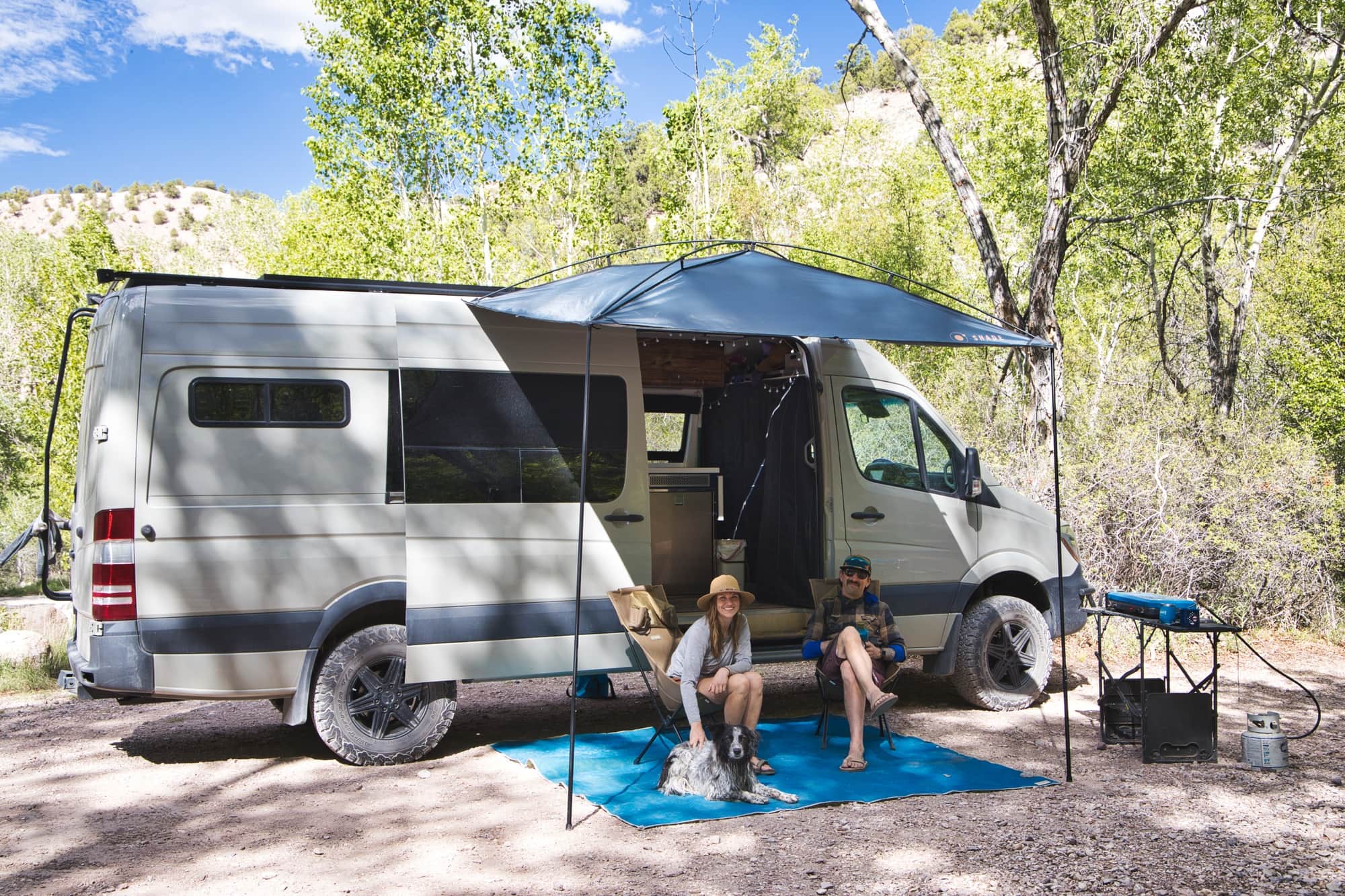 A couple sits with their dog under a van awning at a sunlit mountain campsite.