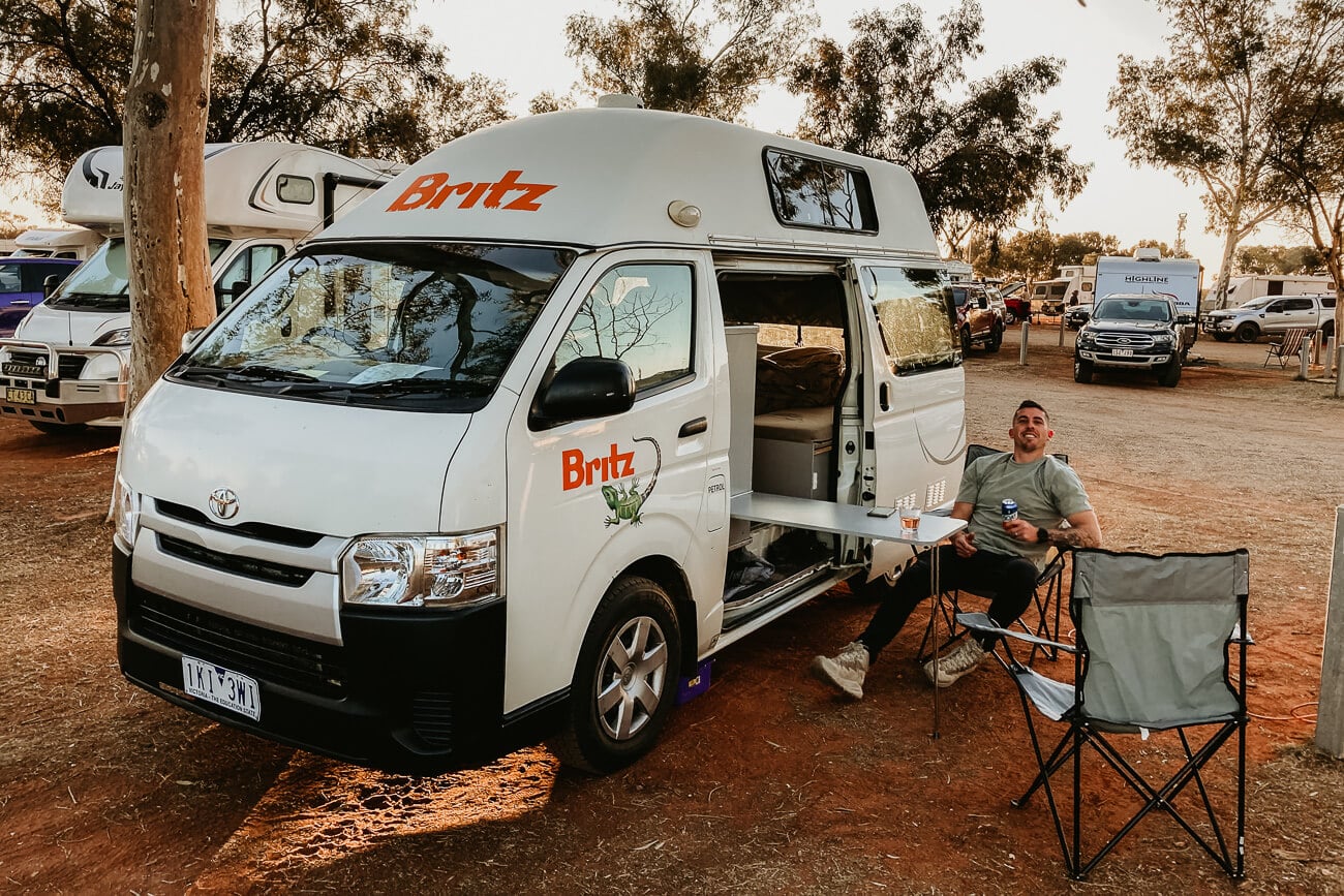 A man sits in a folding chair next to a white Britz campervan at an unpaved campsite during the day.